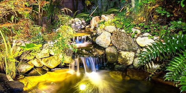 A stunning outdoor scene with a beautifully illuminated pond surrounded by lush greenery. The pond is adorned with underwater lights, casting a mesmerizing glow on the water's surface. In the background, a gentle waterfall cascades down rocks, accentuated by strategically placed spotlights. The image captures the magical ambiance created by DIY landscape lighting for ponds and waterfalls.